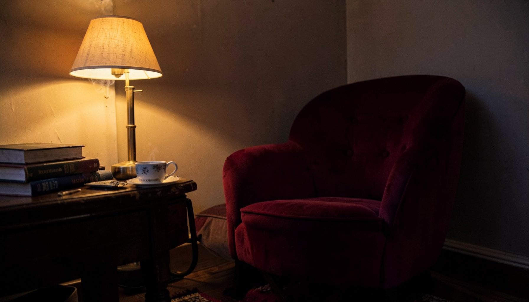 Un rincón acogedor con una butaca de terciopelo granate, una lámpara de luz cálida y libros apilados sobre una mesa de madera.